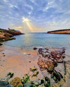 Fotografía de autor de un atardecer en La Caleta de Adeje, Tenerife, con rayos de sol filtrándose entre las nubes sobre una playa de rocas y arena.
