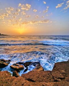 Fotografía de autor del amanecer en la costa de El Médano, Tenerife, con el sol bajo iluminando las nubes y el oleaje rompiendo con fuerza sobre las rocas en primer plano.