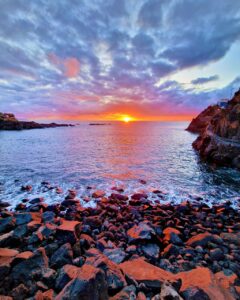 Atardecer sobre una costa de rocas volcánicas oscuras con el sol tocando el horizonte y nubes iluminadas en tonos violetas y naranjas.
